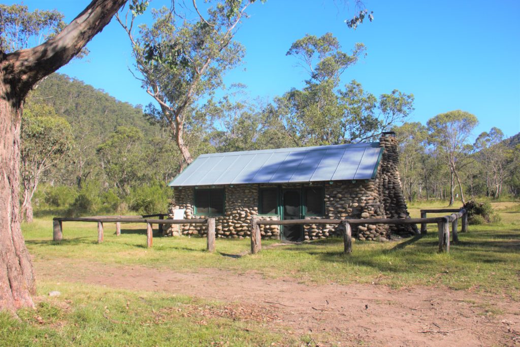 Image shows hut made of stone and mortar, surrounded by wooden fence and trees